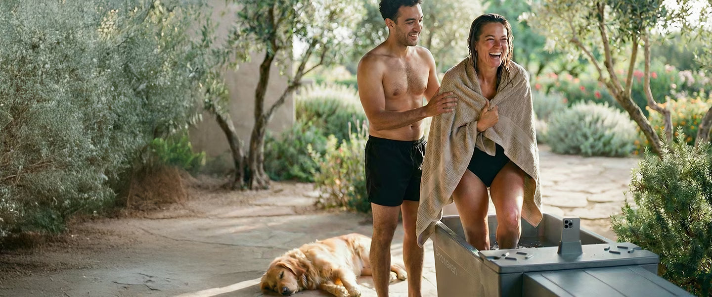 Man and woman standing outdoors with a dog, wrapped in a towel.